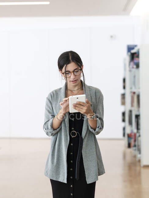 Mujer en gafas usando tableta mientras está de pie en un lugar de trabajo espacioso y trabajando en el proyecto - foto de stock