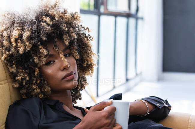 Спокойная афроамериканка с африканскими волосами сидит с кружкой напитка на мягком диване в гостиной и охлаждения в выходные дни дома — стоковое фото