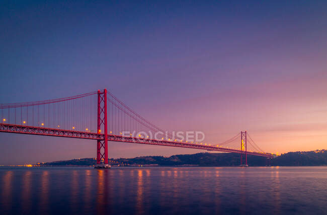 Famous 25 de Abril Bridge crossing Tagus River and connecting Lisbon and Almada near Sanctuary of Christ the King monument against cloudy sundown sky in Portugal — Stock Photo