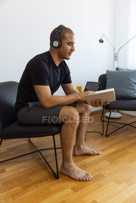 Thoughtful focused male reading interesting book while sitting on chair in living room in morning at home with headphones — Stock Photo