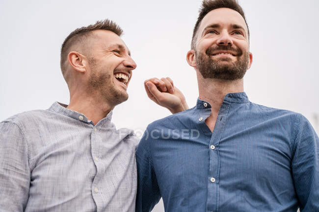 Счастливый мужчина с современной стрижкой смеется, разговаривая с гомосексуальным партнером в рубашке днем — стоковое фото