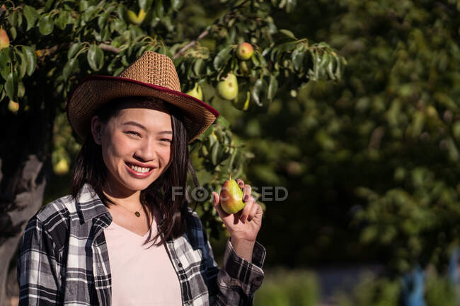 Delighted ethnic female farmer standing with pear in summer garden in countryside and looking at camera — Stock Photo