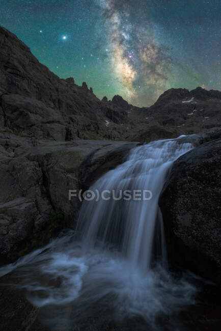 Великолепный пейзаж пенного брызгающего водопада, струящийся по пересеченной скалистой местности под ночным звездным небом с ярким светящимся Млечным Путем — стоковое фото