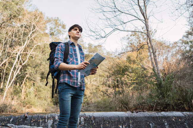 Low angle of male hiker with backpack navigating with paper map while standing in woods and looking away — Stock Photo
