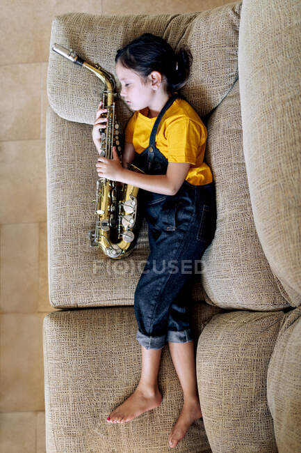 From above side view of barefoot child with saxophone napping on couch in house room — Stock Photo