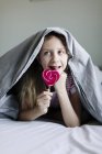 Menina comendo pirulito colorido na cama, foco seletivo — Fotografia de Stock
