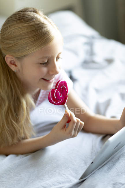Ragazza che utilizza tablet digitale e mangiare lecca-lecca rosa a casa, concentrarsi sul primo piano — Foto stock