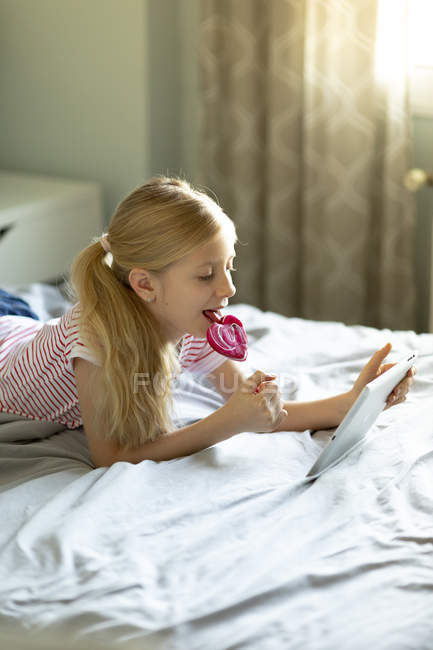 Chica usando tableta digital y comer piruleta en casa, se centran en primer plano - foto de stock