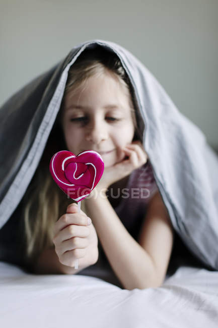 Chica comiendo piruleta de colores en la cama, enfoque selectivo - foto de stock