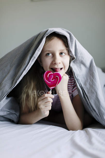 Menina comendo pirulito colorido na cama, foco seletivo — Fotografia de Stock