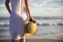 Donna in piedi sulla spiaggia e tenendo il cappello — Foto stock