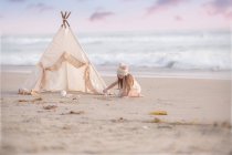 Fille assise par wigwam sur la plage — Photo de stock