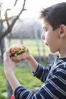 Мальчик держит гамбургер в саду — стоковое фото