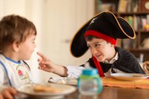Мальчик в пиратском костюме играет с младшим братом, сидящим за столом — стоковое фото