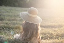 Вид сзади девушки, сидящей в соломенной шляпе в поле — стоковое фото