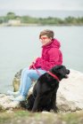 Усміхнена молода дівчина сидить на колоді біля озера з собакою — стокове фото