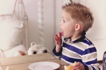 Side view portrait of cute little boy eating sandwich sitting at wooden table with white dog — Stock Photo