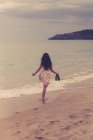 Вид сзади на девушку, идущую вдоль песчаного пляжа с обувью в руке — стоковое фото