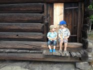 Dos chicos caucásicos con gorras sentados en la escalera del edificio de madera - foto de stock