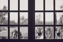 Портрет двух милых игривых маленьких девочек, смотрящих в окно — стоковое фото