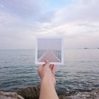 Imagem recortada de mão humana segurando foto de transferência de impressão instantânea em frente ao mar — Fotografia de Stock