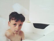 Sério olhando menino sentado no banho com espuma na cabeça — Fotografia de Stock