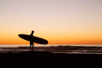 Silhouette eines jungen männlichen Surfers, der mit seinem Surfbrett am Strand in San Diego, Kalifornien, Amerika, USA steht — Stockfoto