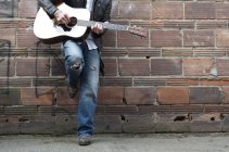 Обрезанное изображение человека в кожаной куртке и ковбойских сапогах, играющего на гитаре в переулке — стоковое фото