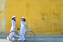 Two women in traditional clothing standing with bicycle on street and talking, Hoi An, Vietnam — Stock Photo
