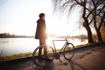 Teenager boy standing with bicycle by river at sunset — Stock Photo