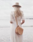 Вид ззаду жінки, що стоїть на пляжі в білій сукні та капелюсі — стокове фото