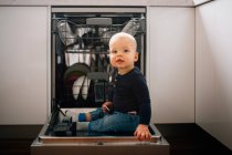 Симпатичный мальчик сидит на открытой двери посудомоечной машины — стоковое фото