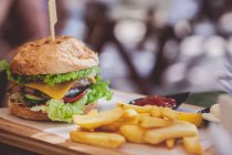 Hamburger and fries on chopping board against blurred background — Stock Photo