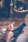 Abgeschnittenes Bild einer Frau in Socken, die zu Hause am Kamin sitzt und eine Zigarette in der Hand hält — Stockfoto