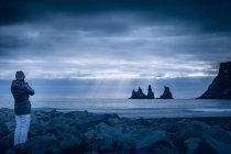 Mujer de pie en el mar sobre nubes tormentosas y mirando a la vista, Islandia - foto de stock