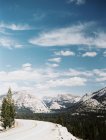 Ciel bleu avec nuages, montagnes enneigées et forêt derrière la route. Route à travers le parc national Yosemite, Amérique, États-Unis — Photo de stock
