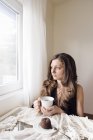 Jovem mulher sentada com xícara de chá e sobremesa de chocolate e olhando através da janela — Fotografia de Stock