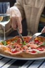 Las manos femeninas cortan un pedazo de deliciosa pizza y un vaso de vino blanco en la mesa - foto de stock