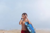 Мальчик стоит на пляже с буги-доской и делает притворный бинокль с пальцами — стоковое фото