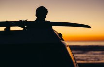 Силует автомобіля і молода людина з дошки для серфінгу на даху проти гарний захід сонця в Сан-Дієго, Америка, США — стокове фото