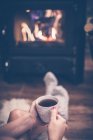 Imagen recortada de la mujer en calcetines sosteniendo la taza de café frente a la chimenea en casa - foto de stock