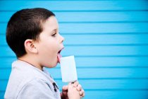 Junge leckt Eislutscher vor blauer Wand — Stockfoto