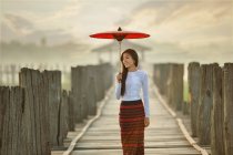 Schöne junge Frau mit rotem traditionellem Regenschirm, die auf einer Brücke steht, Mandalay, Myanmar — Stockfoto