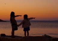 Silhouette zweier junger Mädchen, die bei Sonnenuntergang auf das Meer zeigen — Stockfoto
