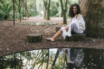 Portrait d'une femme souriante portant une robe blanche assise dans la forêt — Photo de stock