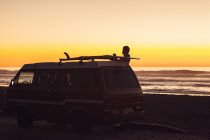 Man putting surfboard on roof rack at sunset on beach — Stock Photo