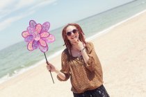 Femme souriante avec dreadlocks debout sur la plage et tenant jouet de moulin à vent de paon en plastique — Photo de stock
