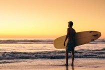 Человек стоит на пляже на рассвете держа доску для серфинга — стоковое фото