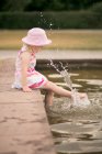 Girl wearing summer dress and pink hat splashing water with feet — Stock Photo