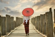 Schöne junge Frau mit rotem traditionellem Regenschirm, die auf einer Brücke steht, Mandalay, Myanmar — Stockfoto